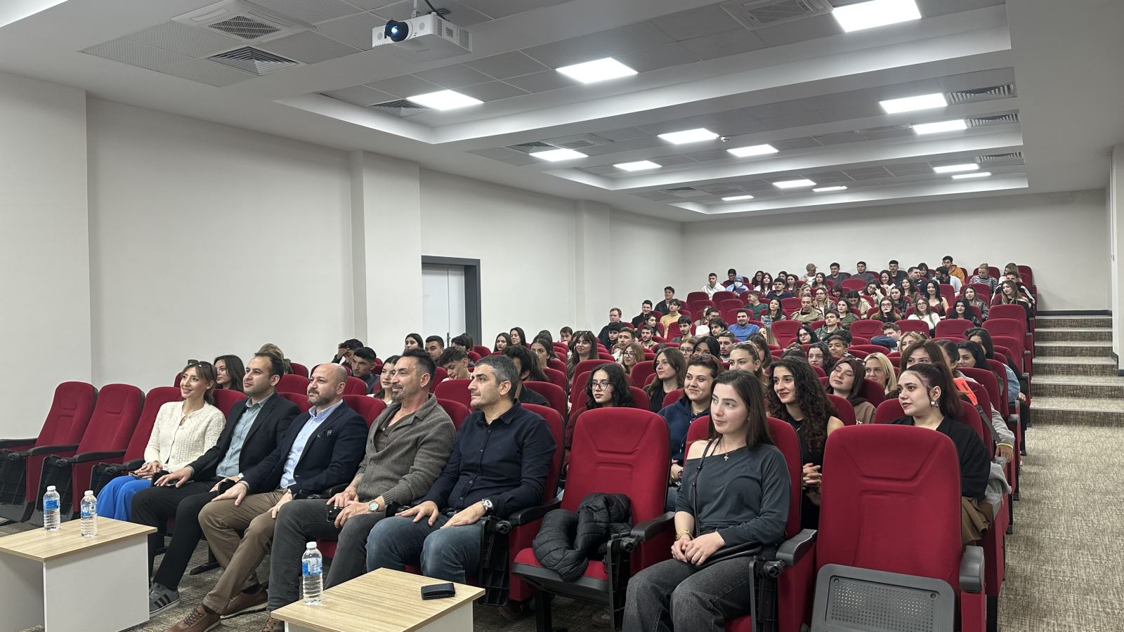 A crowded group of students is seated in red chairs within a large amphitheater. Faculty members and guests are positioned in the front row. The setting is modern and well-lit, with participants focused on the stage.